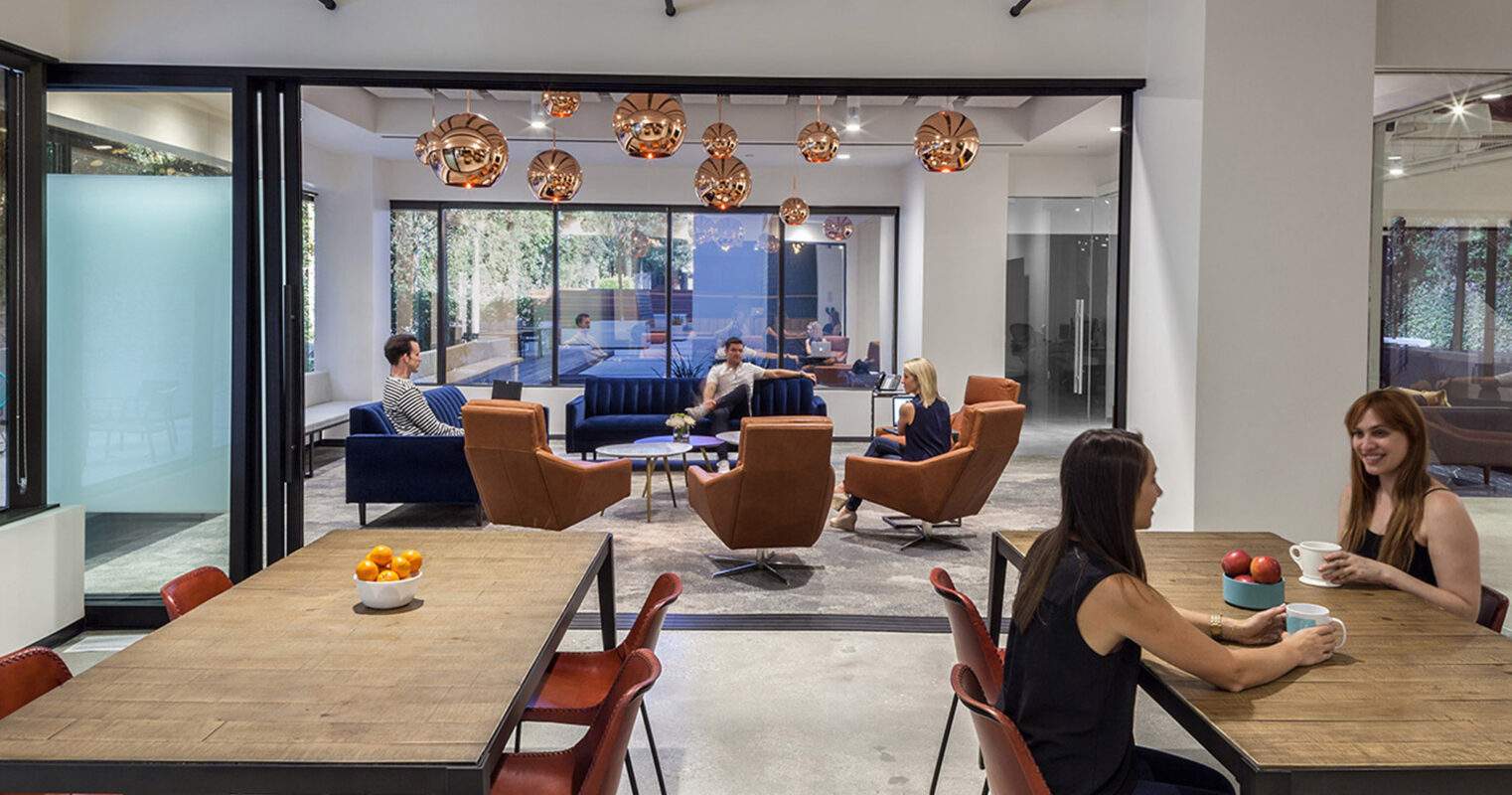 Open-plan office space featuring a harmonious blend of industrial and modern design elements, with polished concrete floors, exposed ductwork, and sleek black window frames. Accentuated by warm-toned copper pendant lighting and rich, upholstered furniture, fostering a collaborative atmosphere.