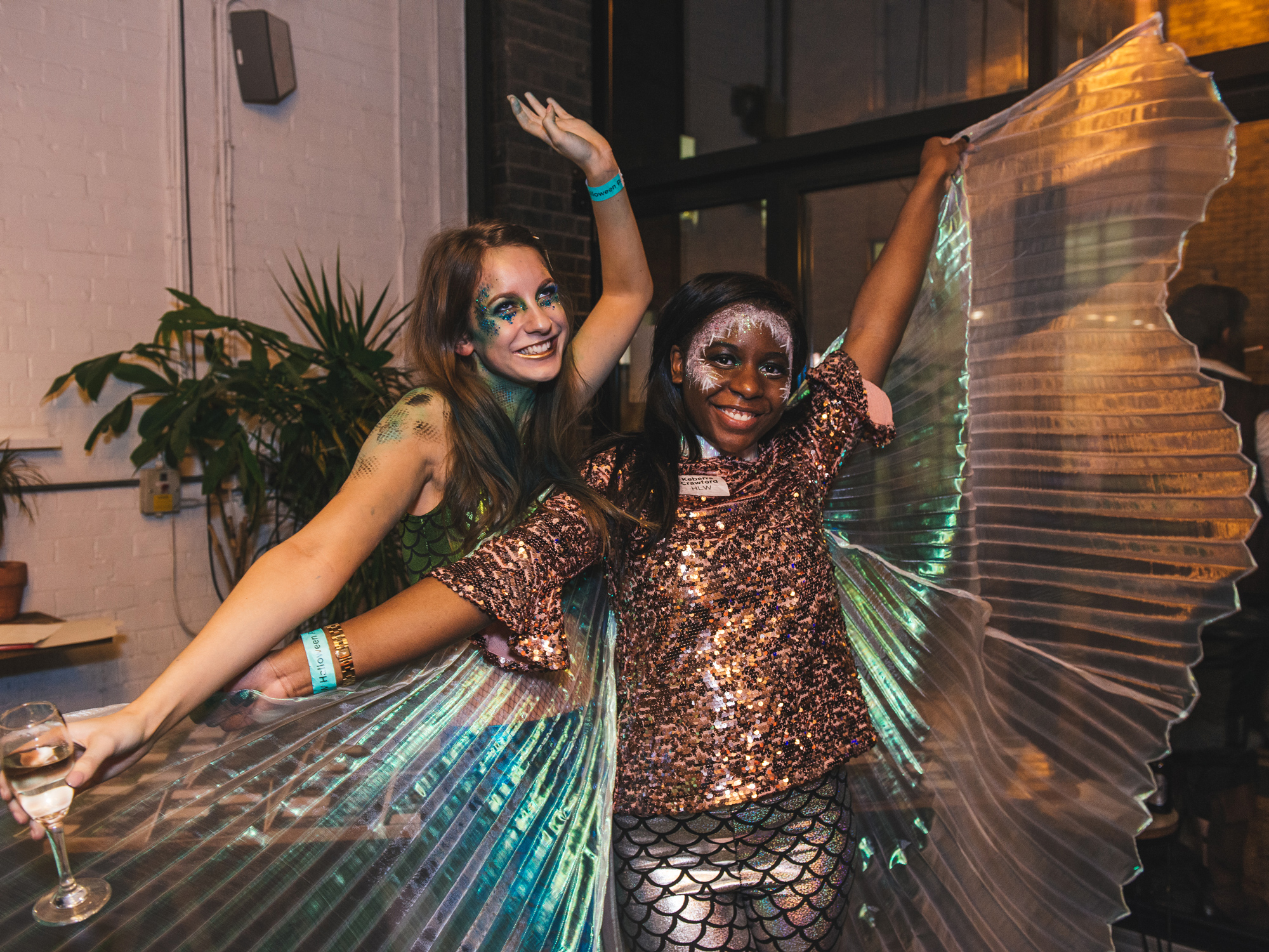 Two individuals in mermaid-inspired costumes with one sporting large, iridescent wings, at a lively costume event.