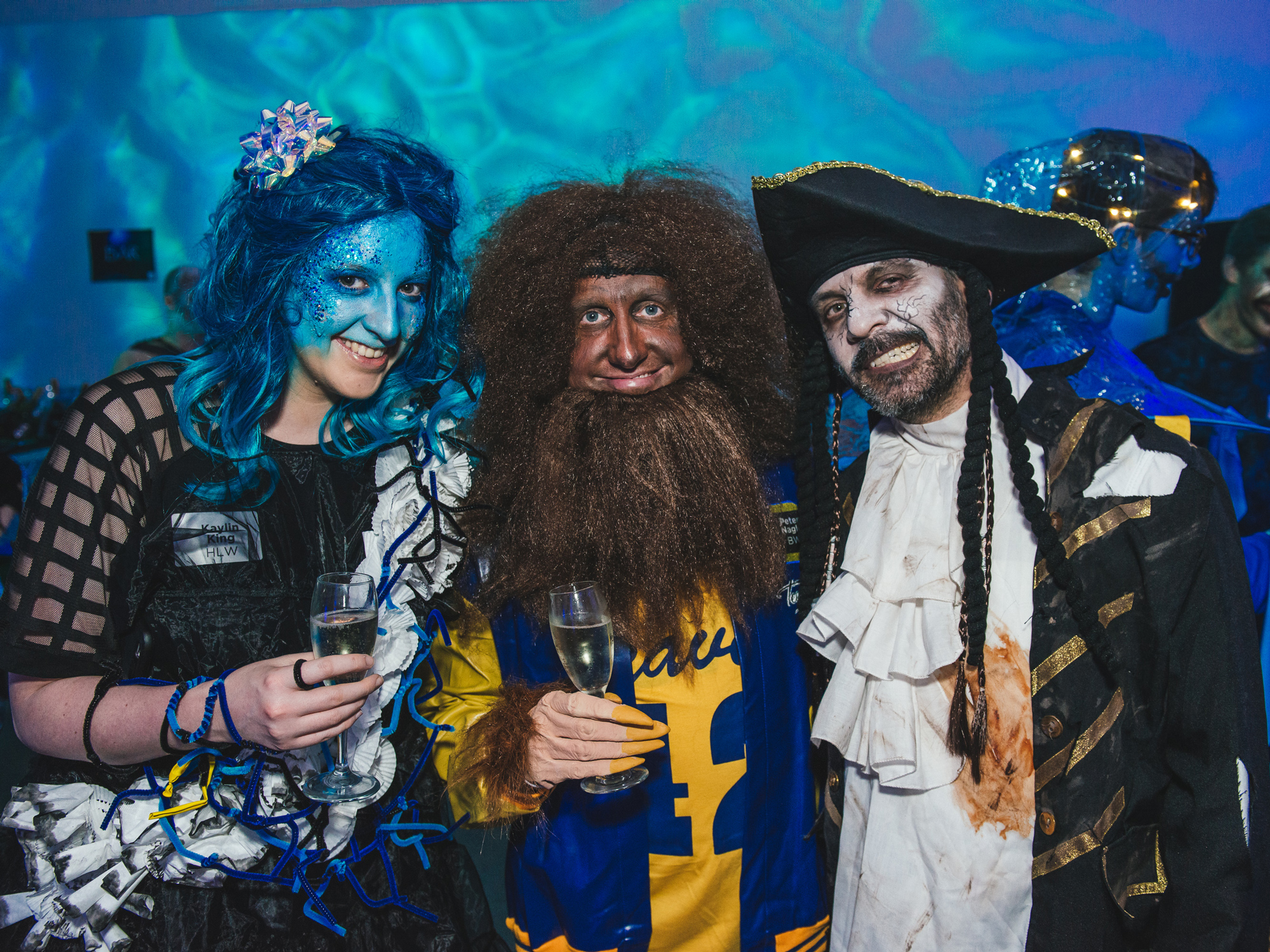 Three costumed attendees in vibrant, mythical creature outfits enjoying a themed event with blue lighting.