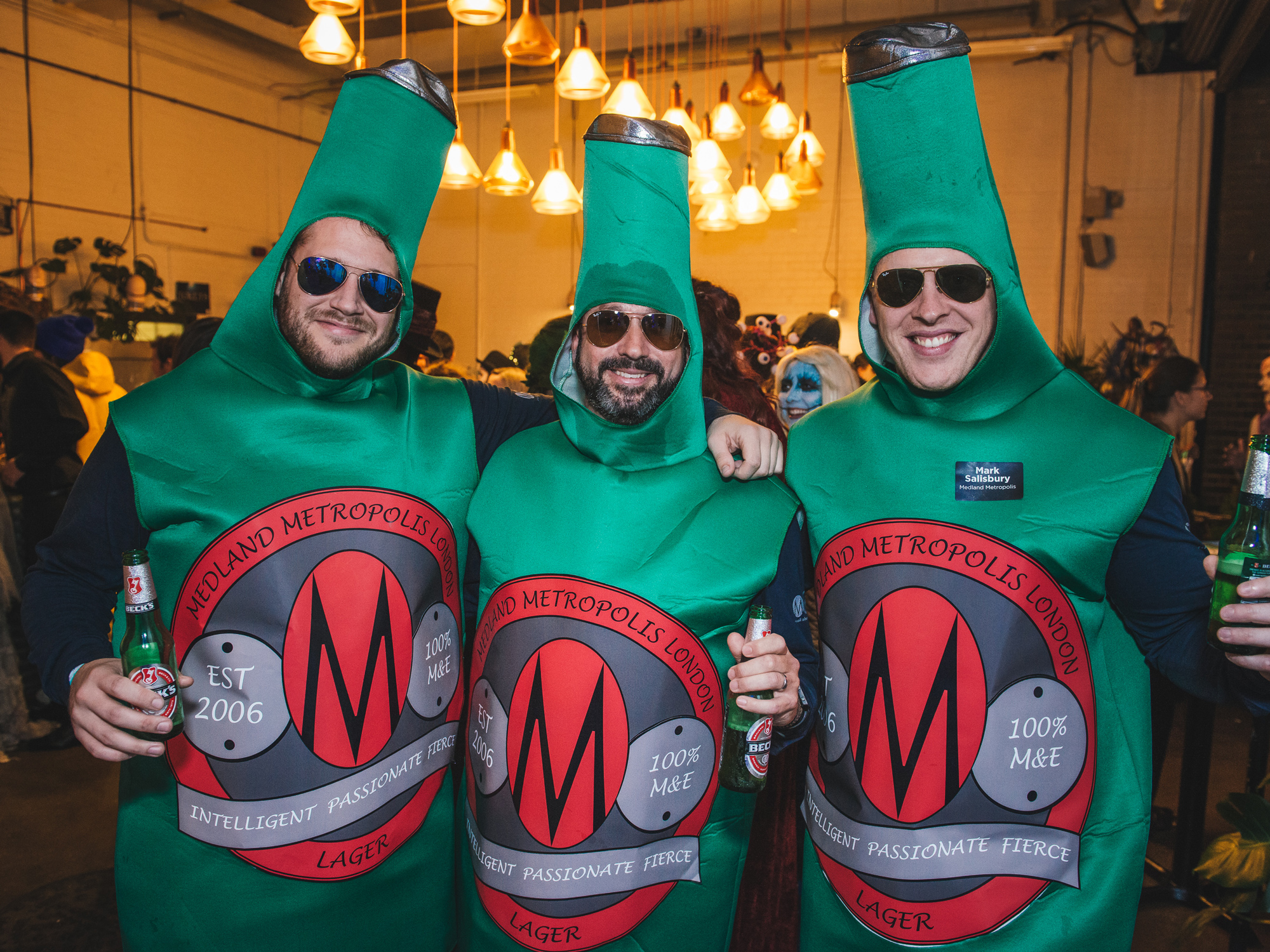 Trio dressed as beer bottles with the Midland Metropolis London logo, enjoying a jovial moment at a social event.