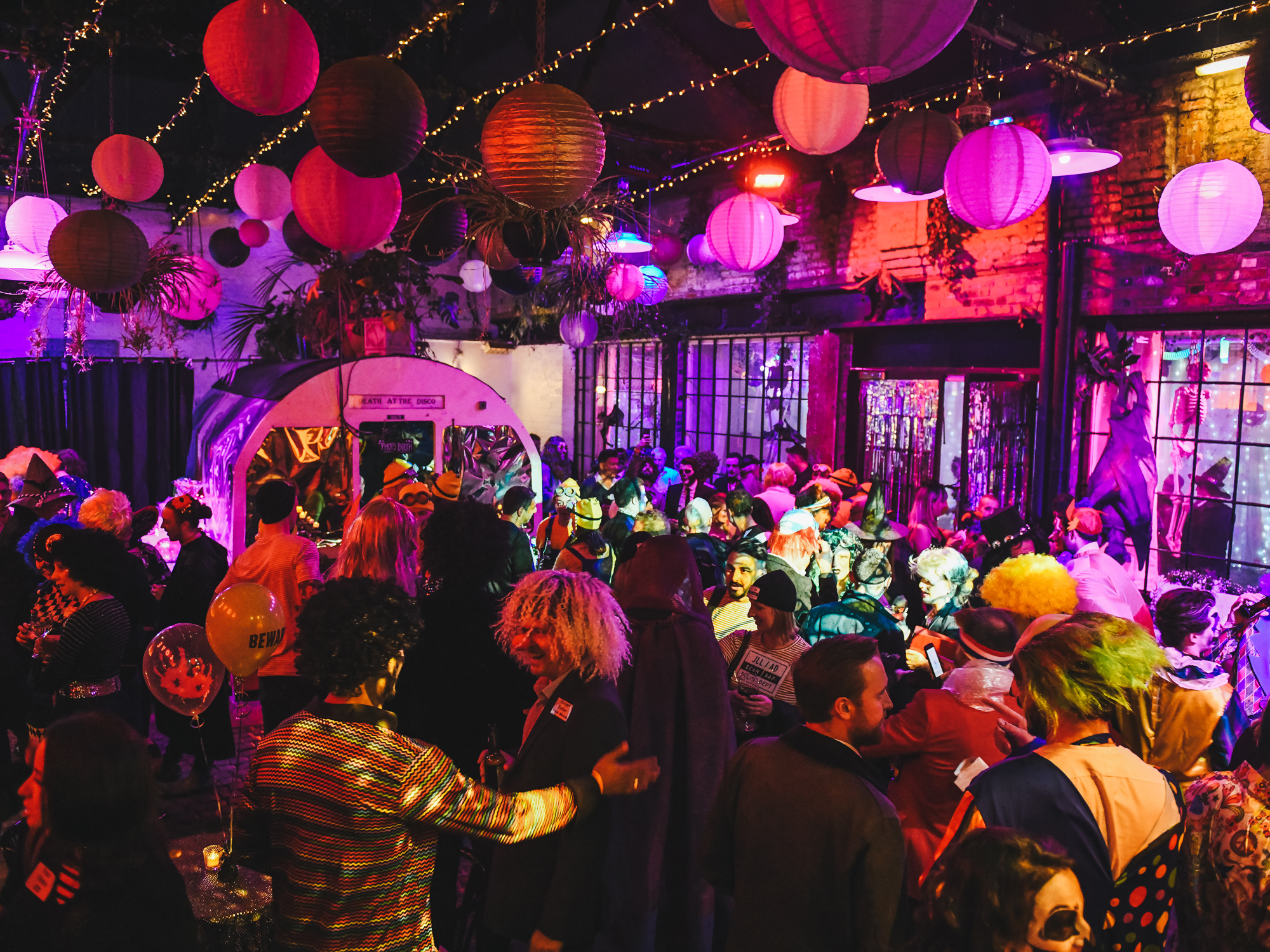 A bustling Halloween party scene with guests in various colorful costumes under decorative paper lanterns.