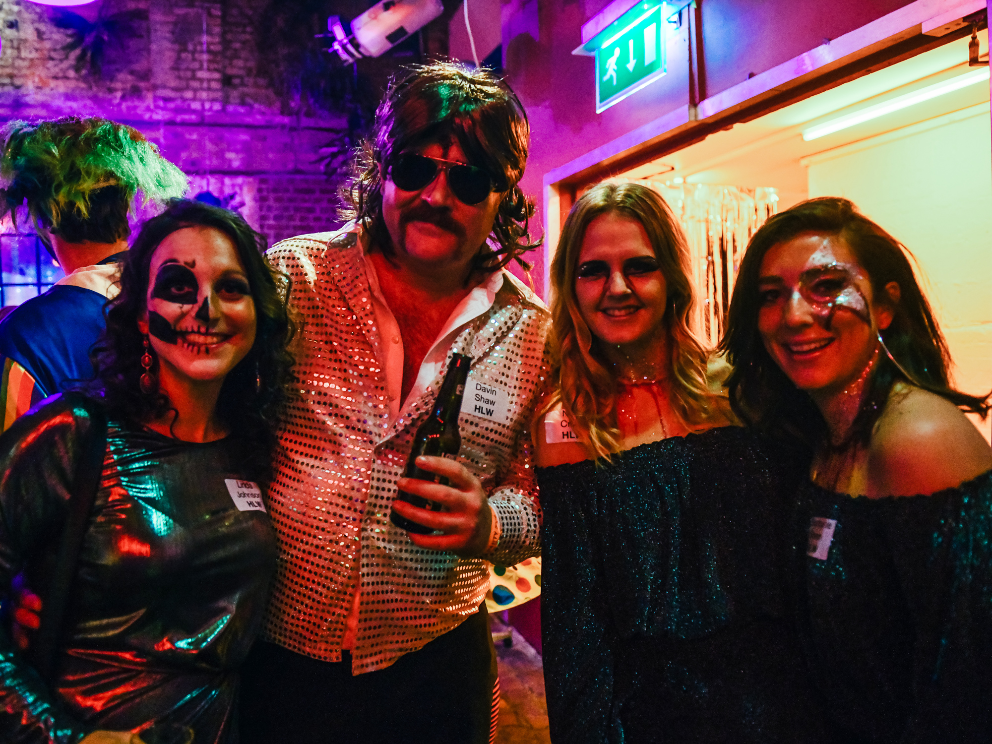 A group of costumed individuals at a Halloween event, one in a bright pink wig, socializing with drinks.