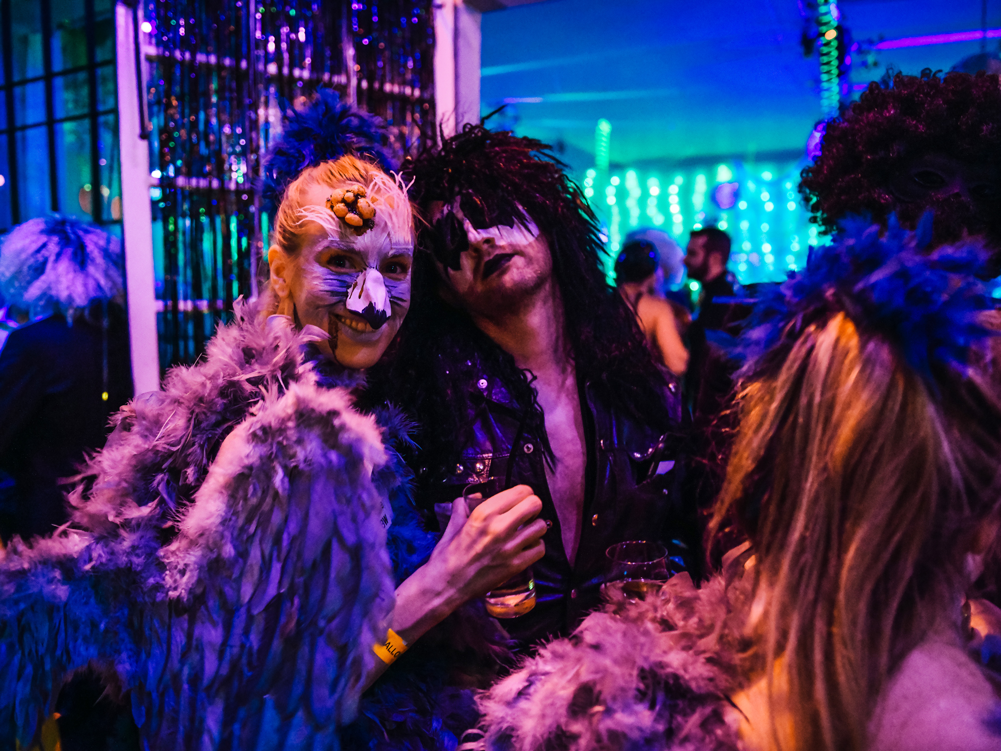 Party-goers in elaborate costumes with feathers and skull makeup, sharing a moment of laughter.