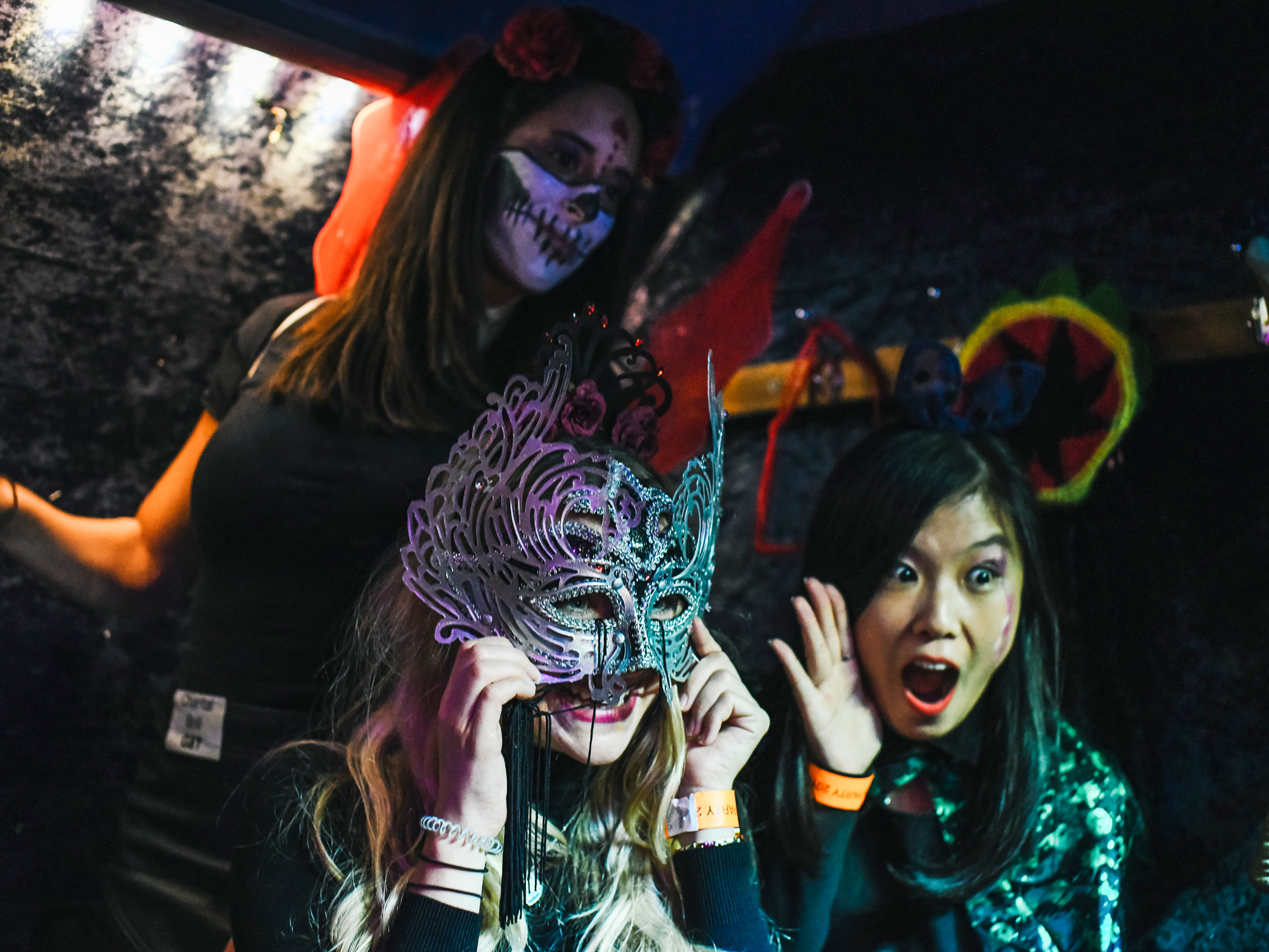 Three women, one in skeletal makeup holding a decorative mask, the other expressing surprise and delight at a party, and the last holding up a mardi gras mask.