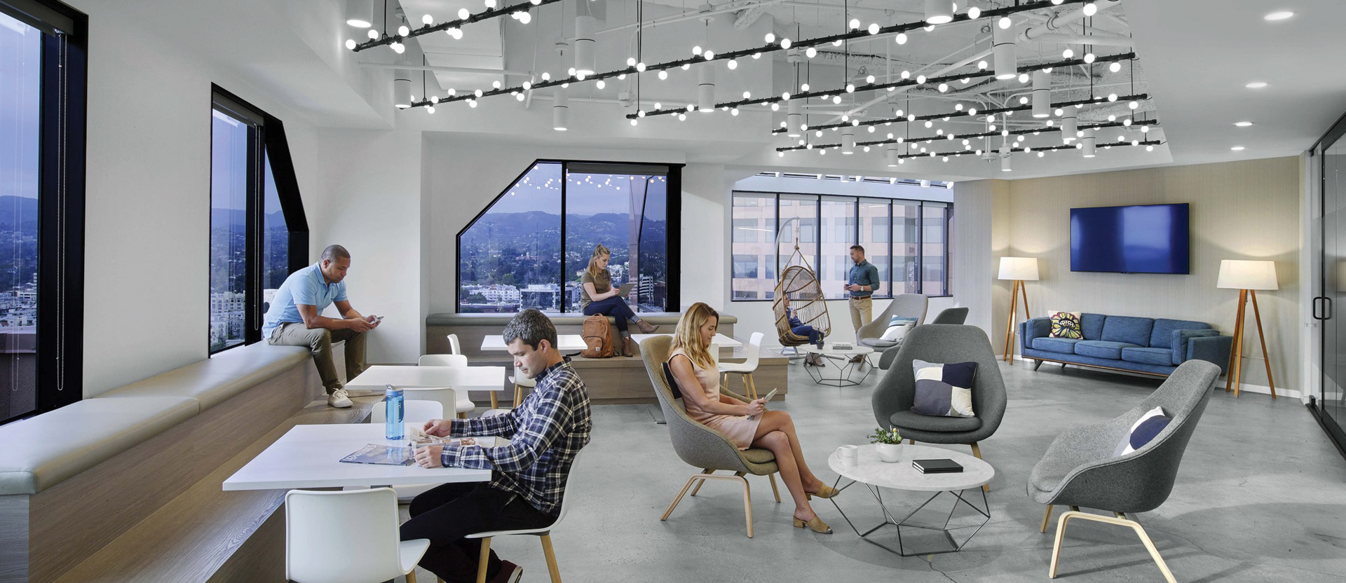 Modern open-plan office space with industrial ceiling, track lighting, exposed ductwork, and floor-to-ceiling windows offering a panoramic view of the cityscape. An eclectic mix of casual seating arrangements includes sofas, armchairs, and collaboration-friendly workstations with employees engaged in various tasks.
