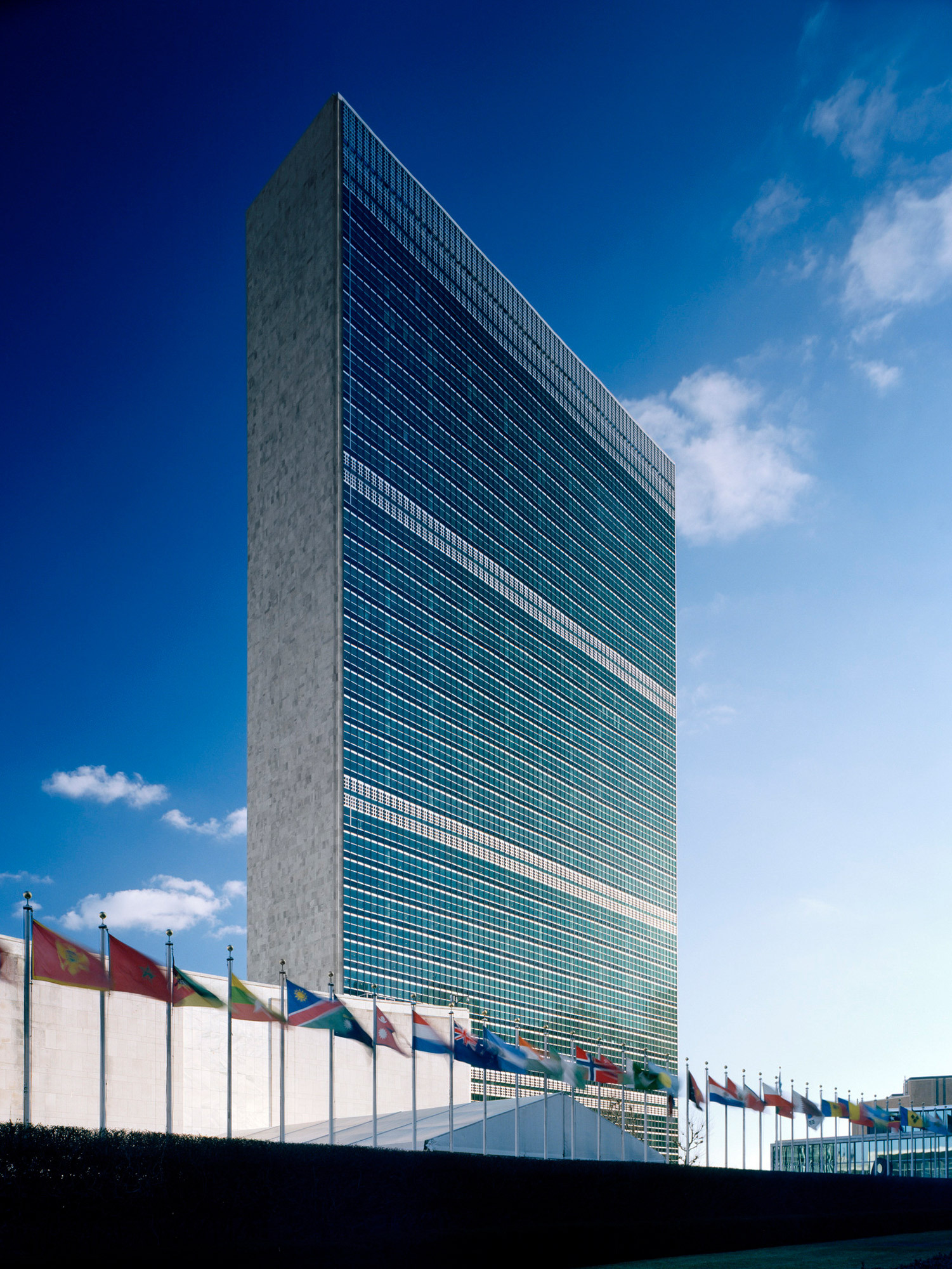 The United Nations Headquarters building stands tall against a clear blue sky, featuring a distinctive grid of windows on its façade, with a row of international flags fluttering in the foreground.