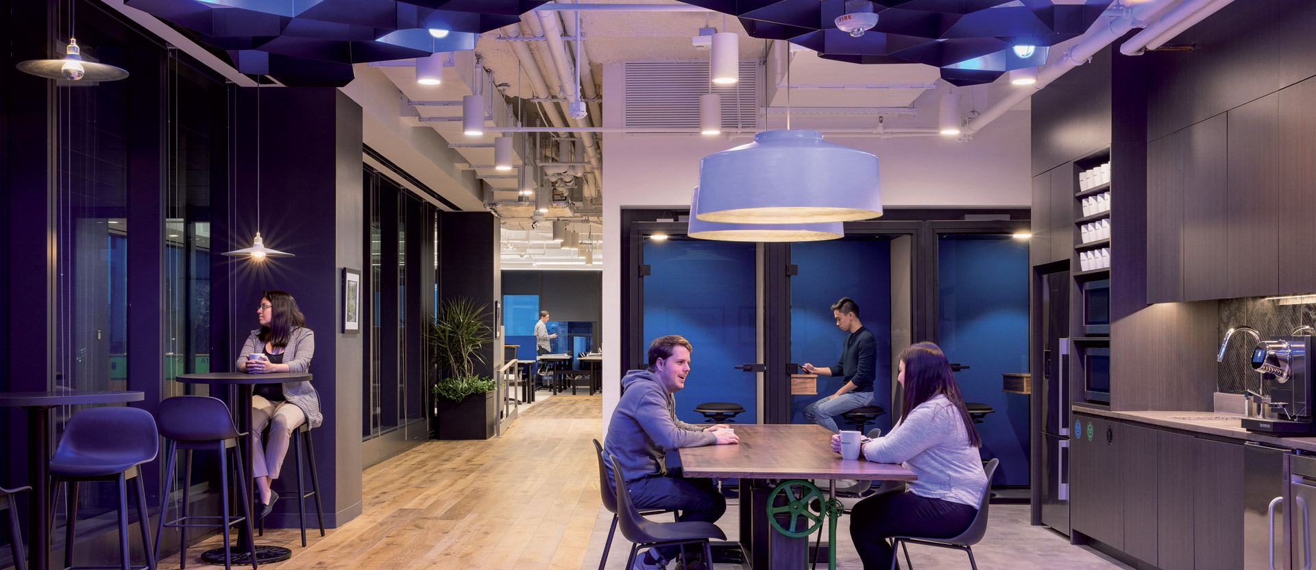 Modern office break room featuring geometric blue acoustic ceiling panels, wooden flooring, and pendant lighting. Colleagues converse at a central table while others work at high-top counters against a kitchenette with dark cabinetry.