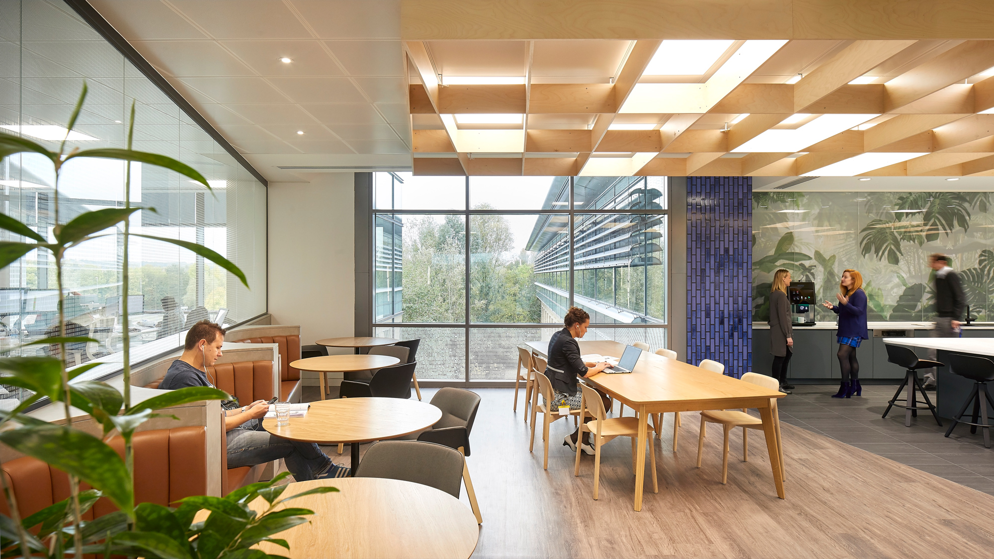 An airy office break room featuring natural wood tables, floor-to-ceiling windows with a view of greenery, and people working and conversing, accented with modern lighting and decor elements.