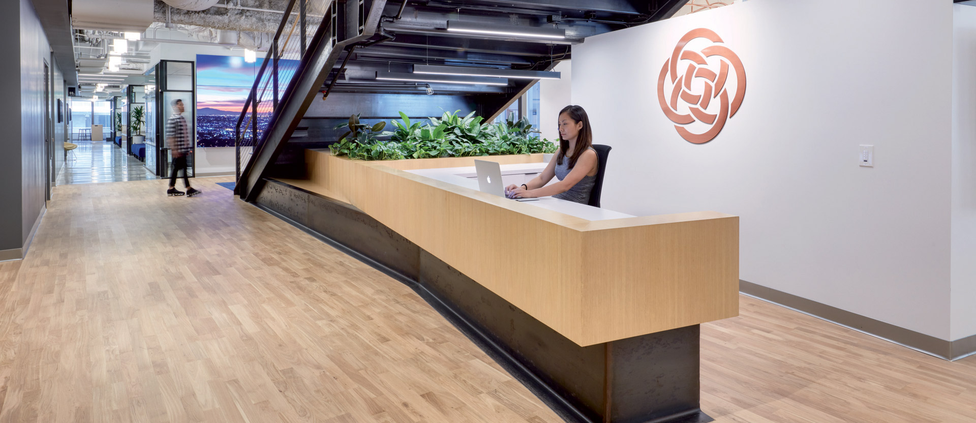 Modern office lobby featuring an angular wooden reception desk with a person at work, live greenery, exposed ceiling ductwork, and sleek metal staircase, creating a balance of industrial and organic design elements.