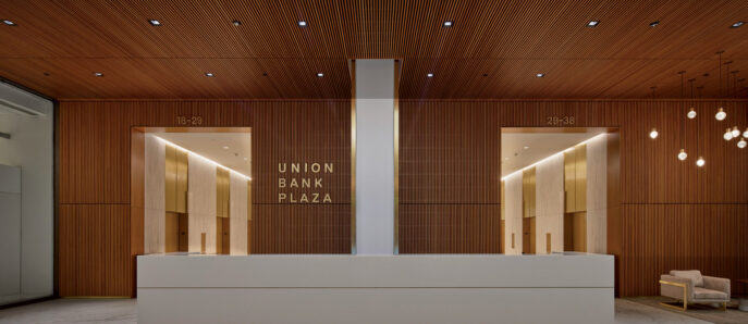 Modern and minimalist reception area featuring a sleek white desk against a textured wooden slat backdrop. Overhead linear lighting accents the warm tones, and recessed floor lighting adds a dramatic effect near the entrance marked "Union Bank Plaza".