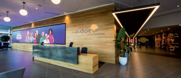 Modern office lobby with a prominent triangular lighting fixture above a sleek reception desk showcasing the Audible logo. Ambient lighting and indoor greenery complement the space, highlighting the brand's corporate identity.