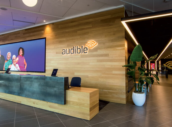 Modern office lobby with a prominent triangular lighting fixture above a sleek reception desk showcasing the Audible logo. Ambient lighting and indoor greenery complement the space, highlighting the brand's corporate identity.