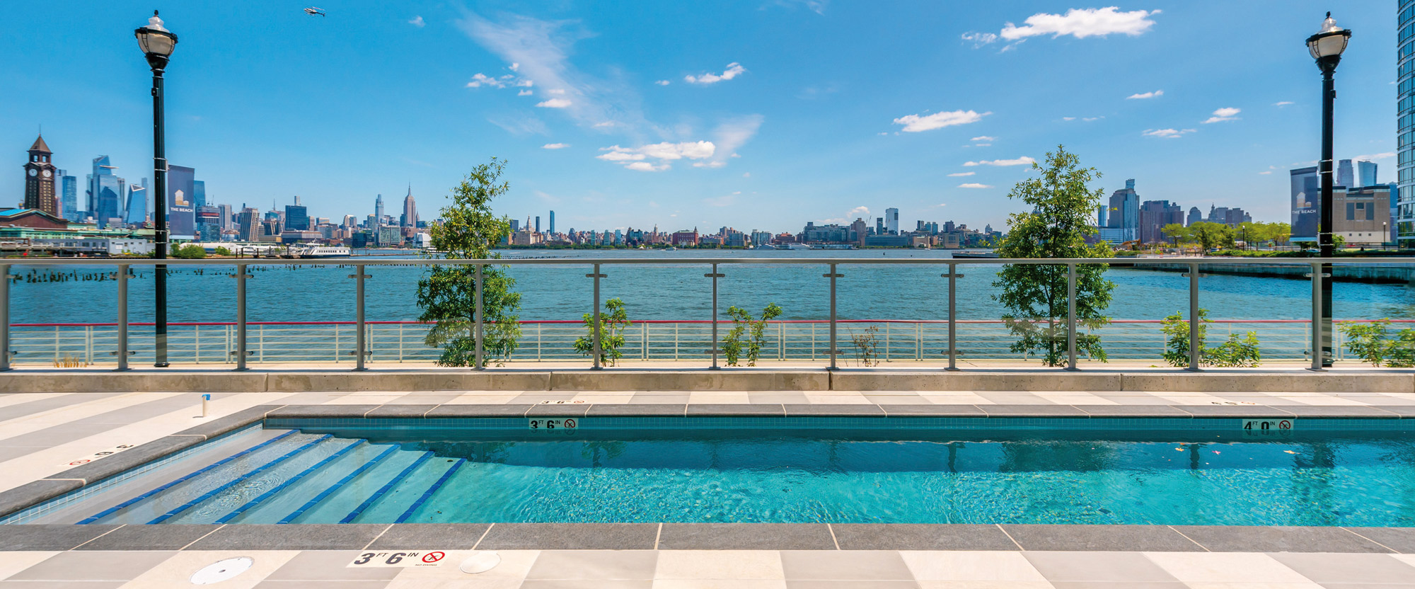 A sleek rooftop pool featuring symmetrical tilework overlooks a scenic city skyline with expansive blue skies and wispy clouds.