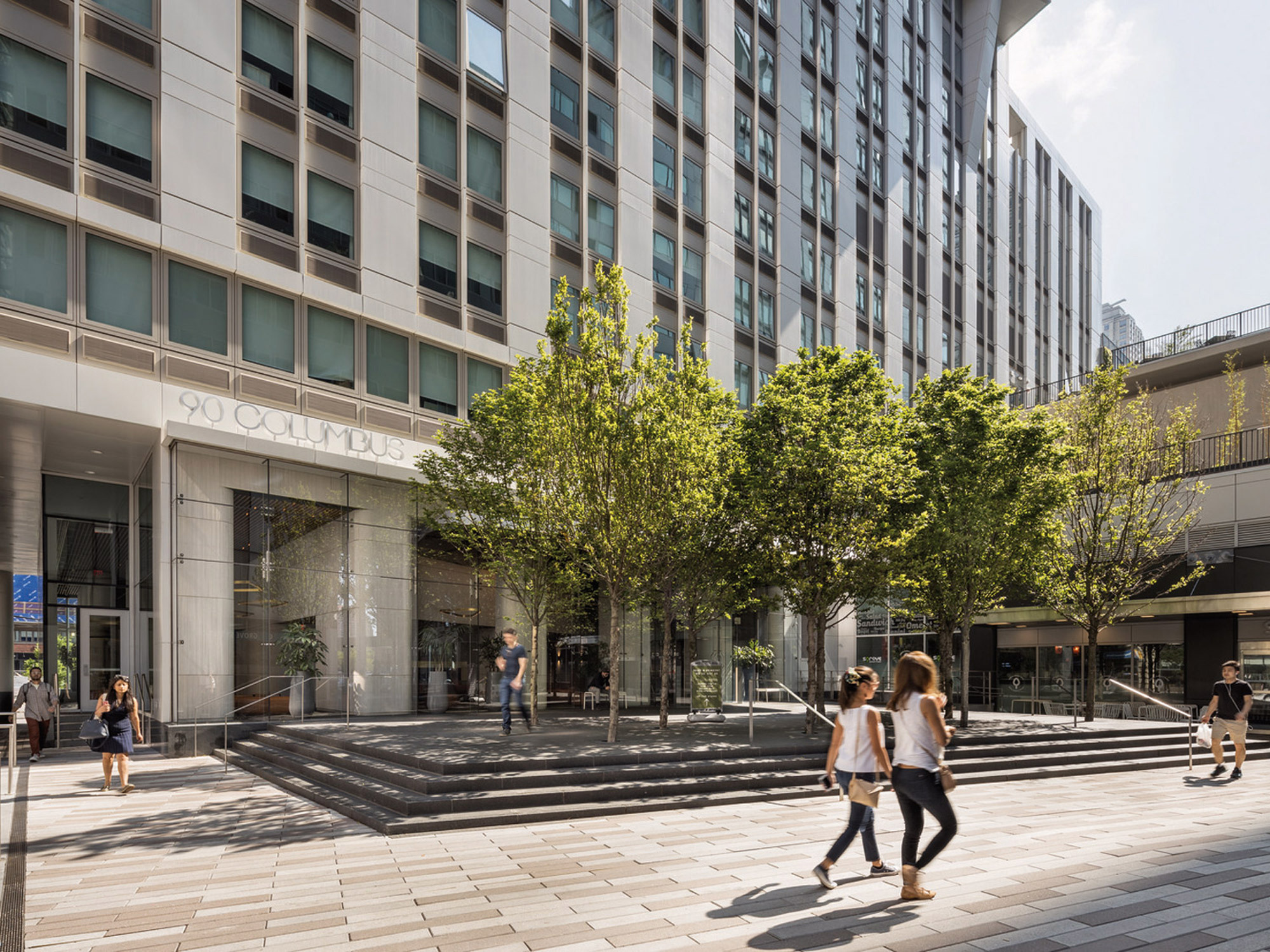 Modern urban streetscape with pedestrians, highlighting a sleek building facade featuring a mix of glass and stone elements, accented by a series of deciduous trees and an inviting entryway marked by a prominent address sign.