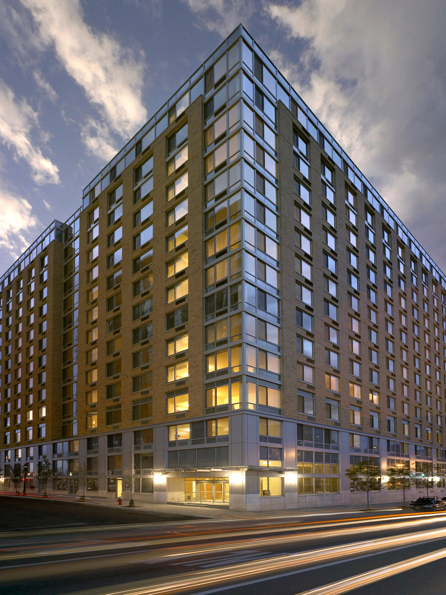 Modern multi-story building captured at dusk, featuring a mix of reflective glass windows and warm-toned brick cladding, with dynamic street-level lighting adding vibrancy to its urban setting.