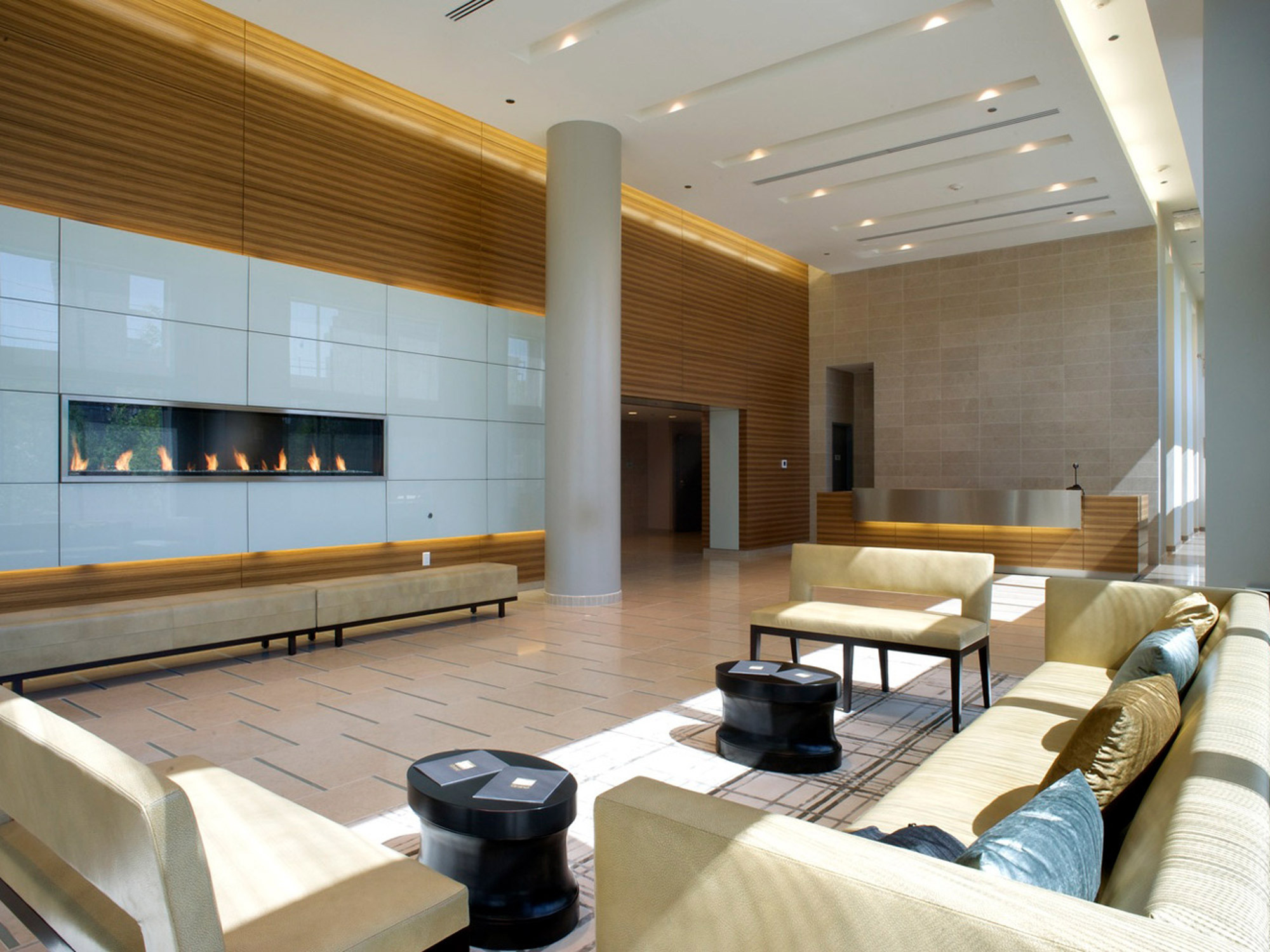 Spacious lobby with floor-to-ceiling windows allowing natural light to accentuate the warm wooden wall panels. A modern fireplace serves as a focal point, flanked by low-profile seating arrangements featuring clean lines and neutral tones, harmonizing with the minimalist aesthetic.