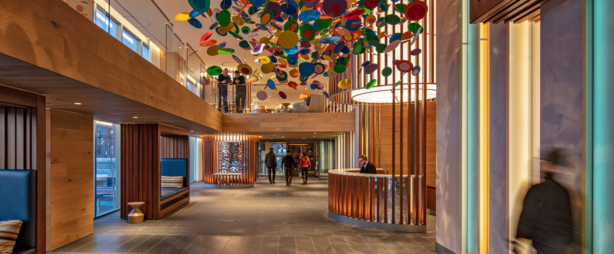 Spacious lobby with soaring ceiling, adorned by a vibrant array of suspended colored orbs, flanked by sleek wooden panels and illuminated recessed wall niches with changing hues, complementing the cool grey floor tiles.