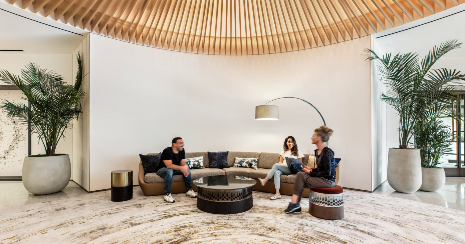 Spacious lounge area with a curved wood slat ceiling radiating from a central point, complemented by minimalist furnishings on a marble floor. Large potted plants add a natural touch to the modern, airy space.