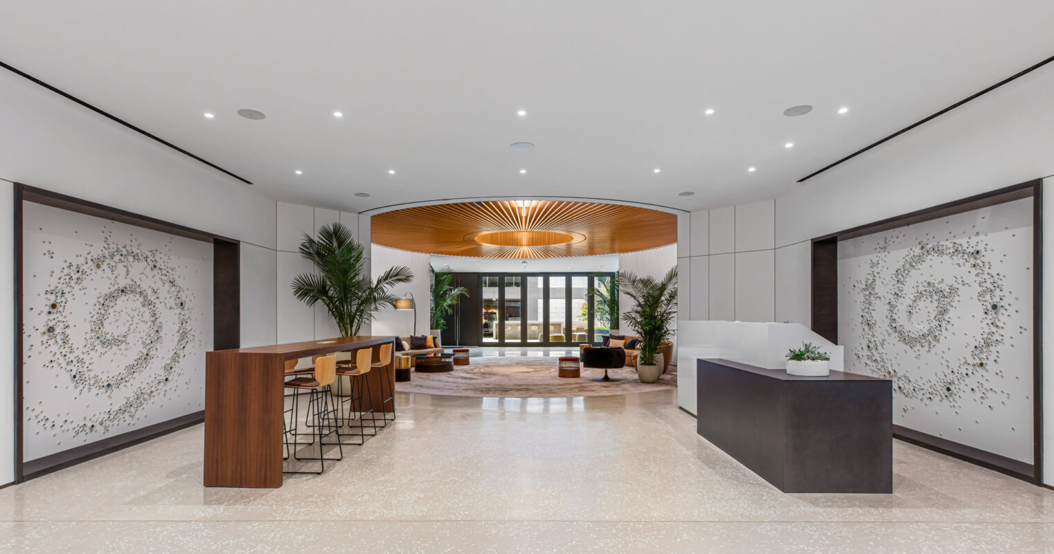 Modern lobby with a sleek marble floor, featuring a wooden curved ceiling accent that guides the eye towards glass doors. Symmetrical decorative panels flank the entrance, with minimalistic furniture subtly enhancing the open and elegant space.