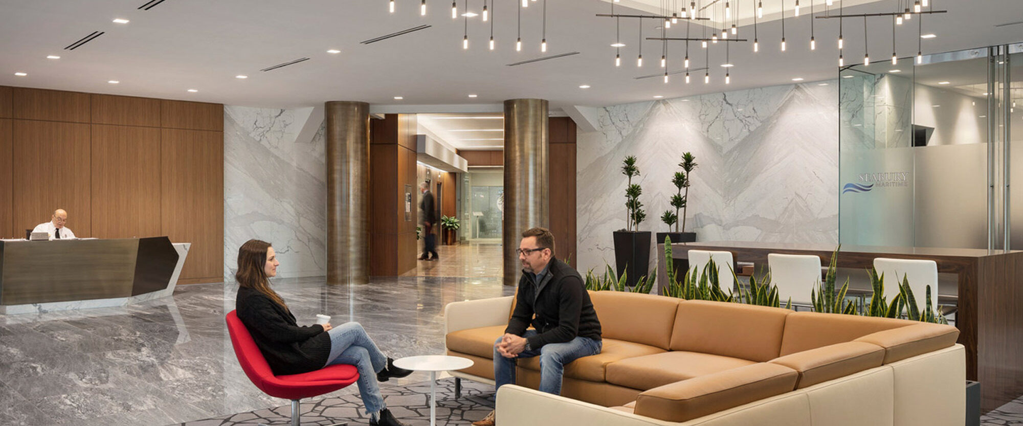 Modern lobby with geometric marble flooring, neutral-toned modular seating, and a reception desk under an array of suspended linear light fixtures. Lush green plant accents complement the sleek, inviting space.