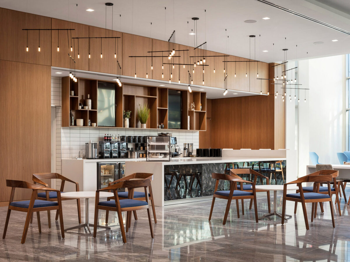 Modern café interior featuring clean lines, warm wood tones on shelving and furniture, with pendant lighting creating a geometric pattern above. The space balances natural materials and sleek design, complemented by floor-to-ceiling windows allowing ample natural light.