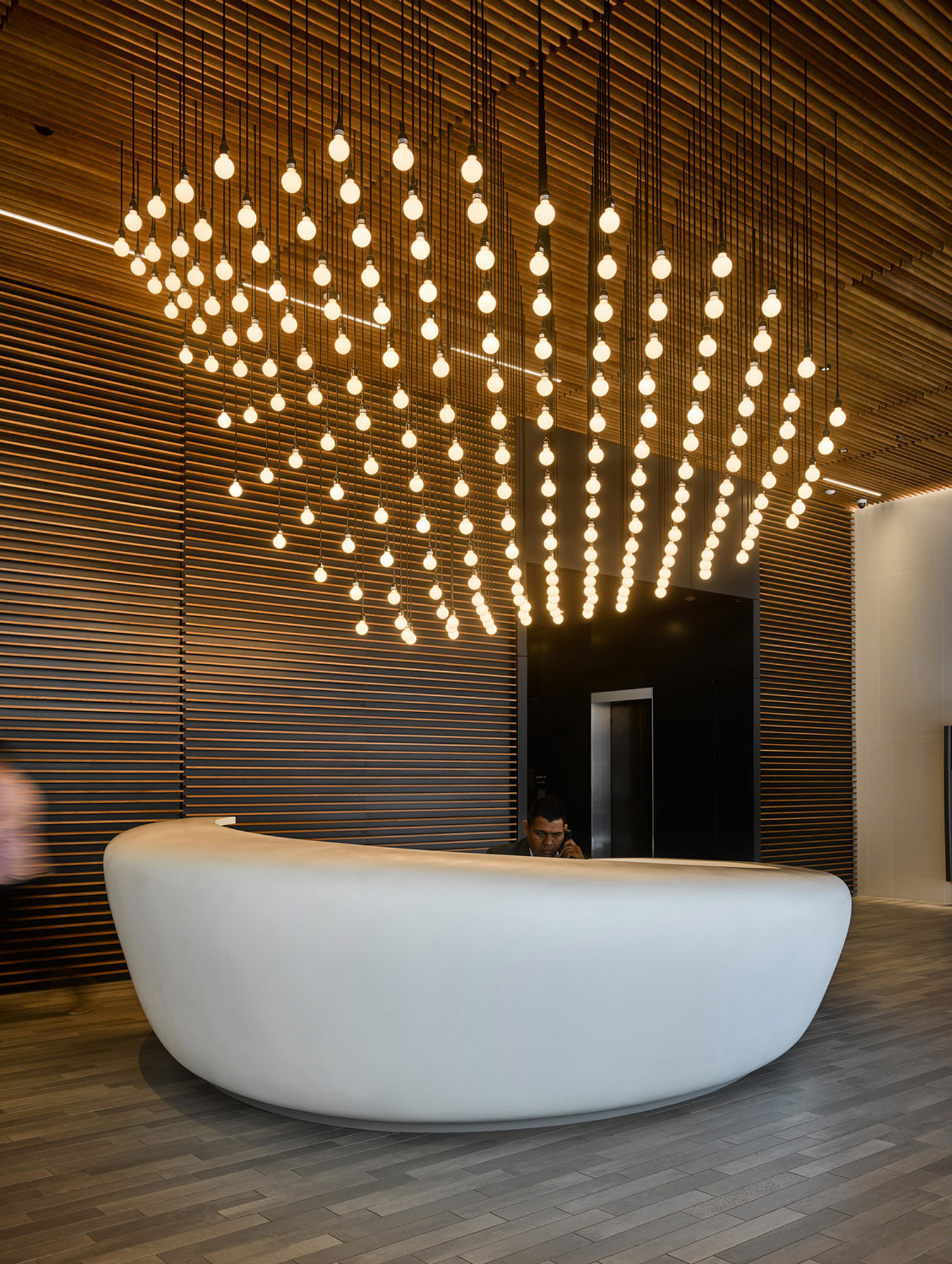 Modern reception area featuring a sleek, curved white desk against a backdrop of vertical wooden slats. Overhead, a striking array of suspended cylindrical lights forms a dynamic, cascading chandelier, enhancing the space with warmth and a contemporary aesthetic.