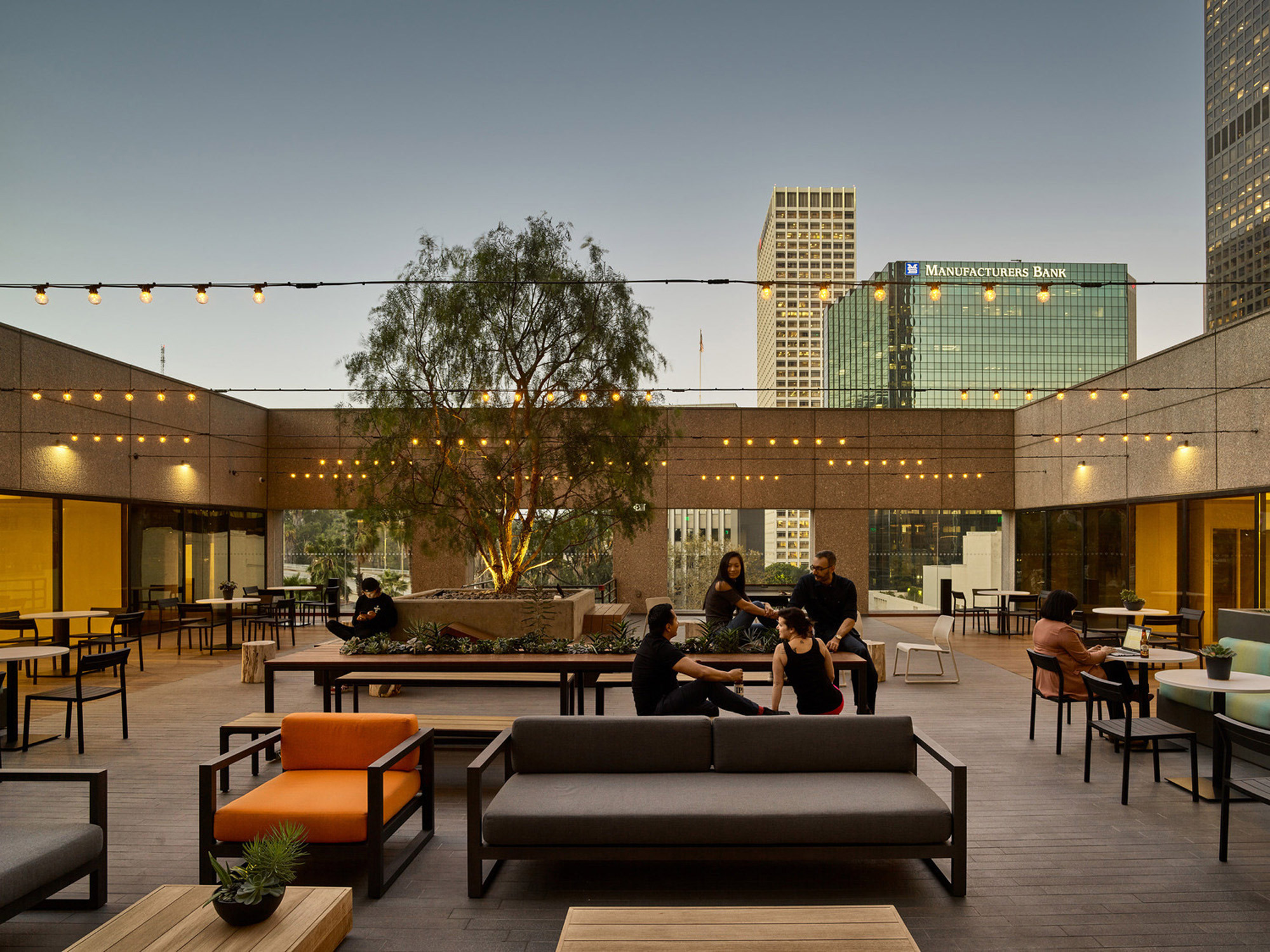 Rooftop terrace with modern outdoor furniture, string lights overhead, and cityscape backdrop during twilight. The area features a mix of communal tables, lounge seating, and landscaping, promoting social interaction in an urban setting.