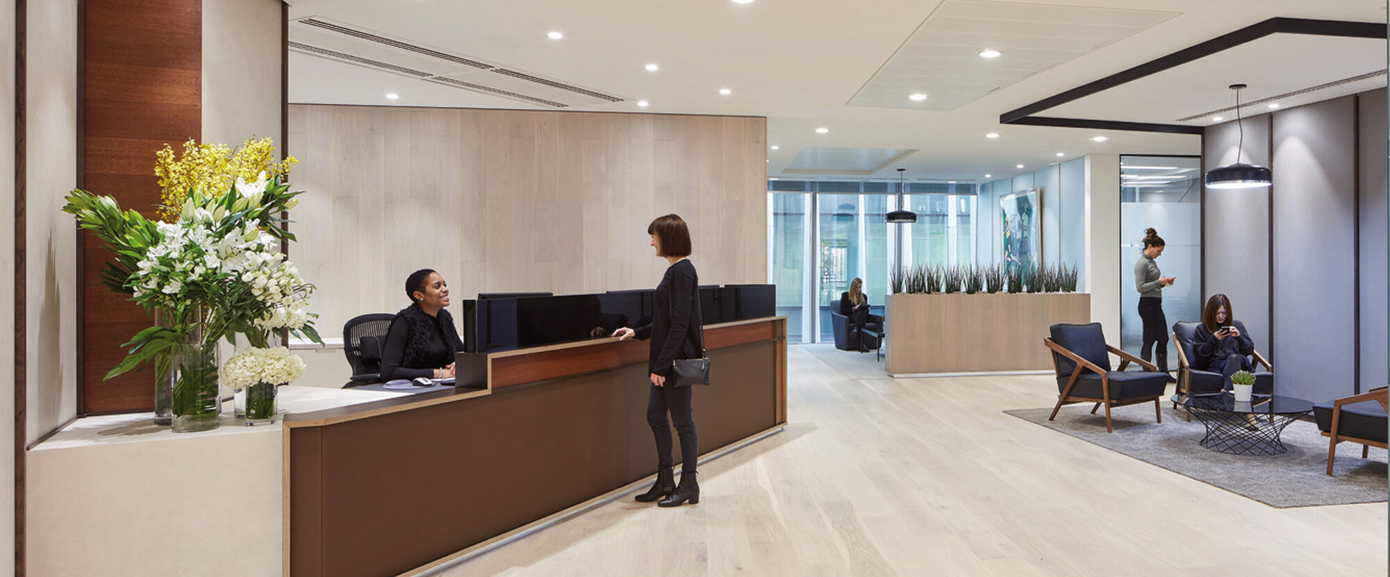 Modern office lobby with light wood floors, a sleek reception desk, and a casual seating area. Neutral tones with natural light give the space a welcoming ambience. Contemporary art pieces adorn the walls, enhancing the professional yet comfortable environment.