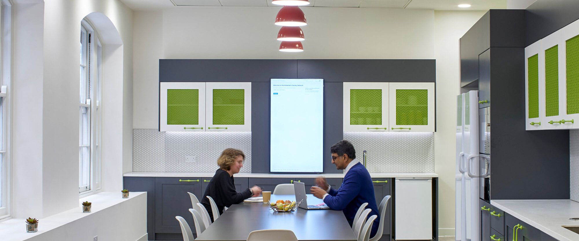 Spacious modern office kitchenette with white walls featuring green soundproofing panels. Slate grey cabinetry and appliances complement the bright red pendant lights. Two professionals engage in conversation at a sleek, central dining table with designer wire chairs.