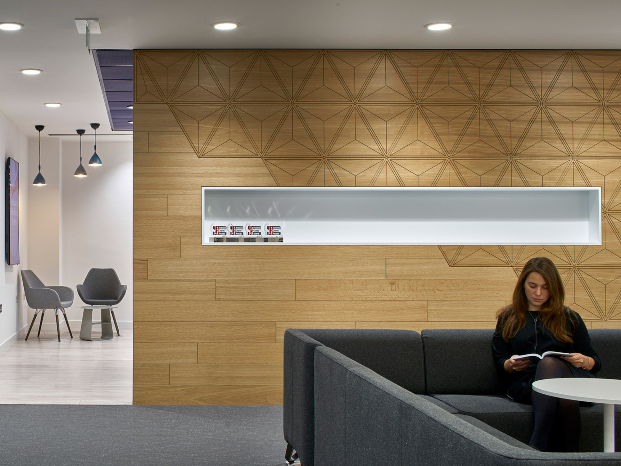 Modern office reception area featuring a geometric wood panel wall, minimalist pendant lighting, and a comfortable seating arrangement with a woman leisurely reading a magazine.