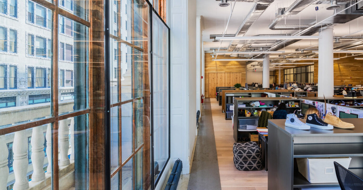 Modern office interior capitalizing on natural light with large, floor-to-ceiling windows flanked by exposed brick. Contemporary workstations with ergonomic chairs punctuate the open-plan space, complemented by warm wooden floors and a view of the urban exterior.