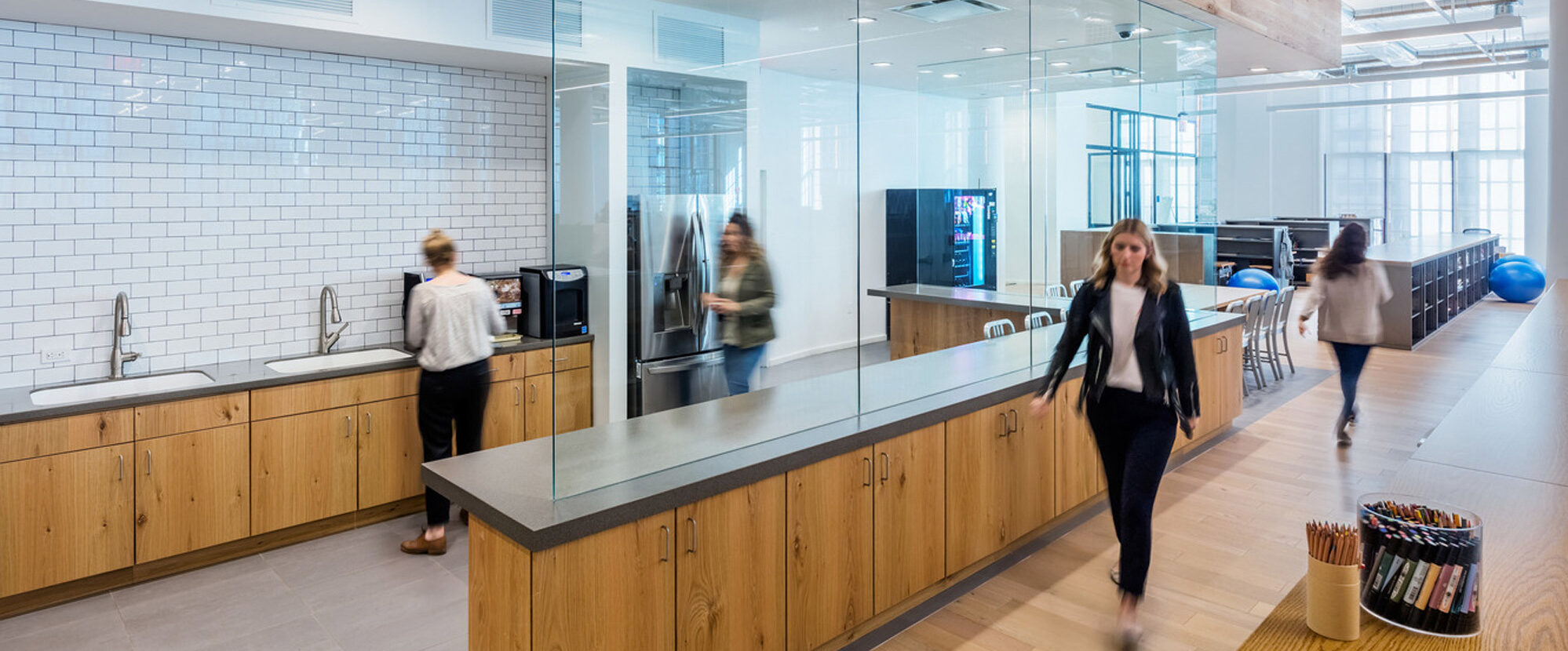 Modern office kitchenette with glass partitions, featuring wood grain lower cabinets against white subway tiles. Exposed wooden beams add warmth, complementing the polished concrete floor. Openness and natural lighting enhance the space's collaborative atmosphere.