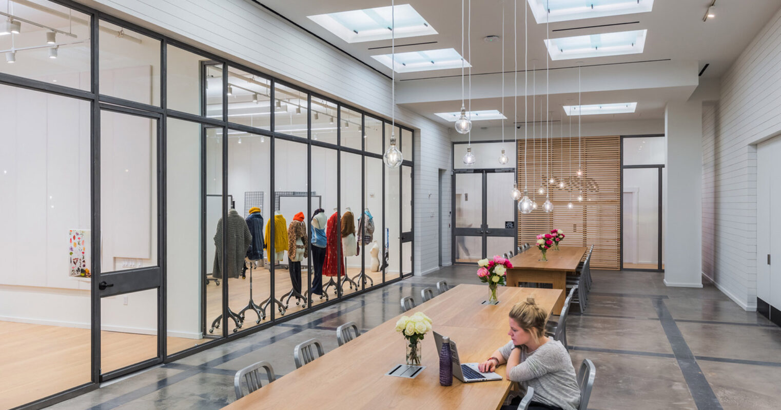 Modern open-plan retail space with skylights, featuring a central communal table, sleek black-framed glass partitions, and clothing racks against an exposed brick accent wall. Vibrant floral arrangements add color to the neutral palette.