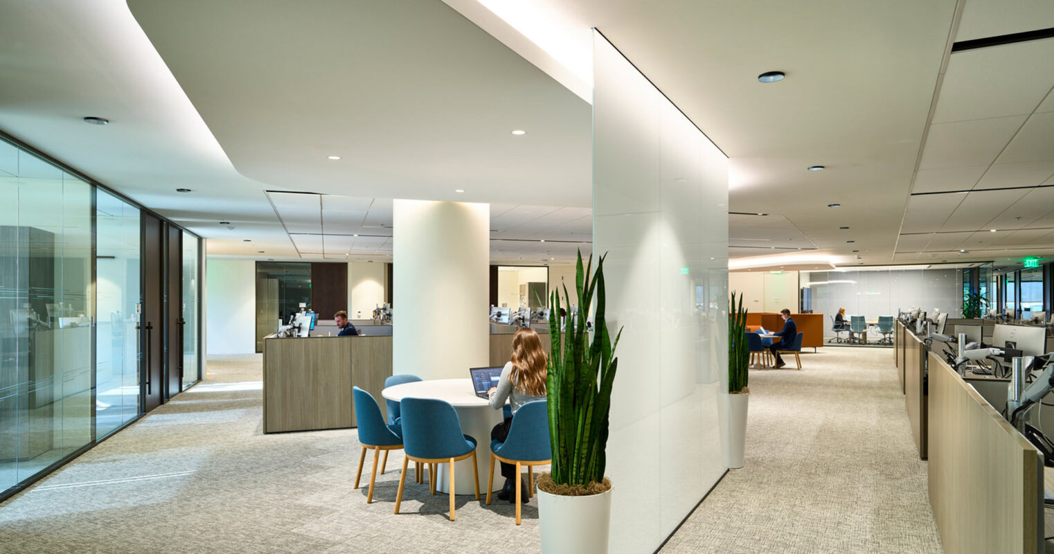 Modern office interior featuring sleek white columns, curvilinear ceiling accents, and textured carpeting. Ambient lighting complements minimalist furniture arrangements while plant accents add a touch of biophilic design.