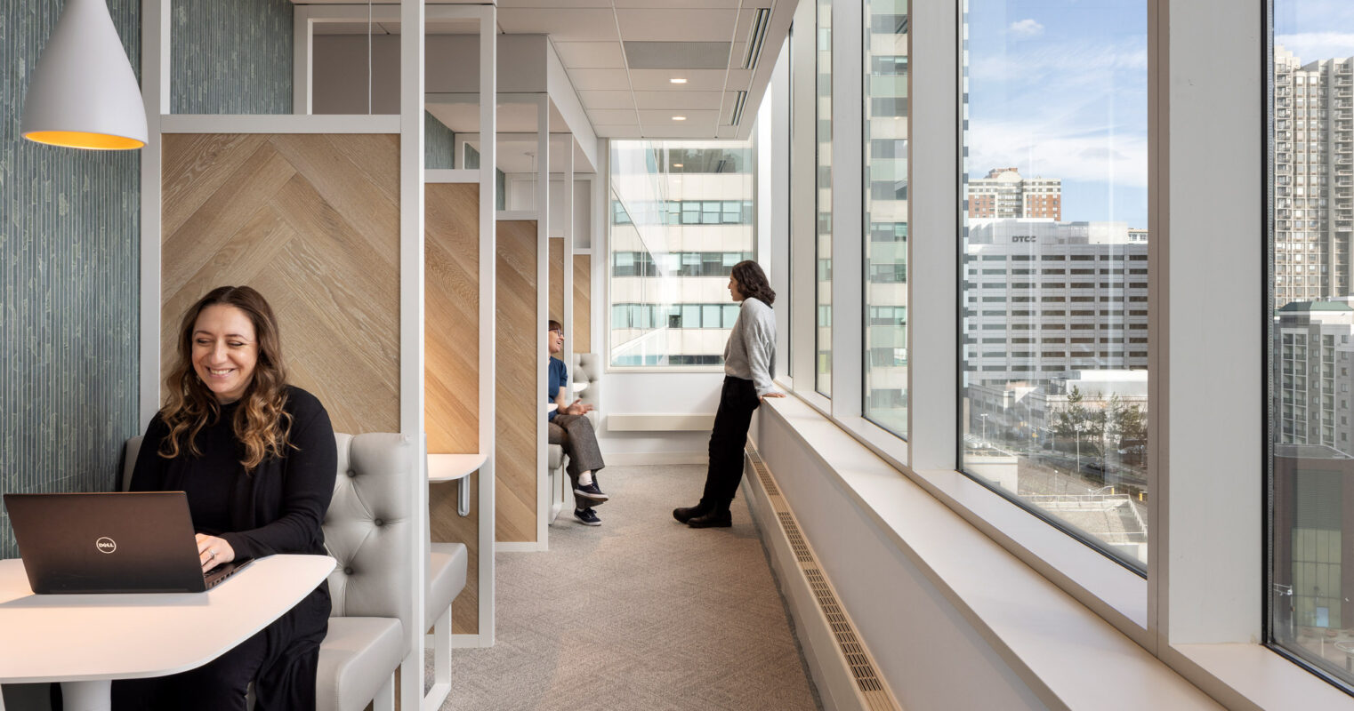 Light-filled office corridor with herringbone wood accents and individual work pods on one side and floor-to-ceiling windows offering a city view on the other, with people engaged in various work activities.