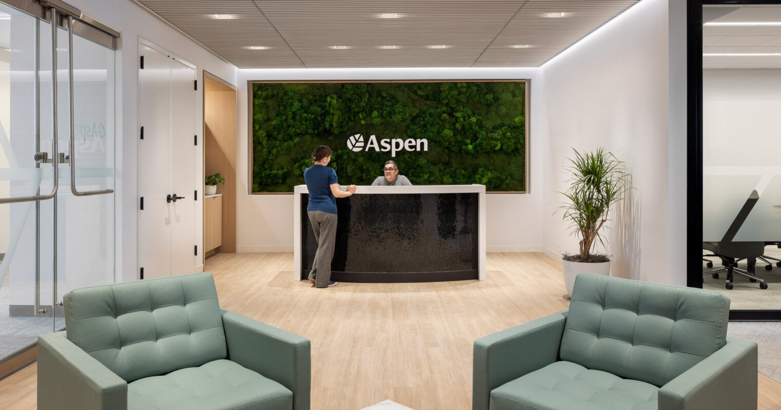 Modern office lobby with a living green wall backdrop, featuring the Aspen logo. Streamlined reception desk with two employees and geometric wood flooring contrast with plush sea-green armchairs and minimalist glass partition.