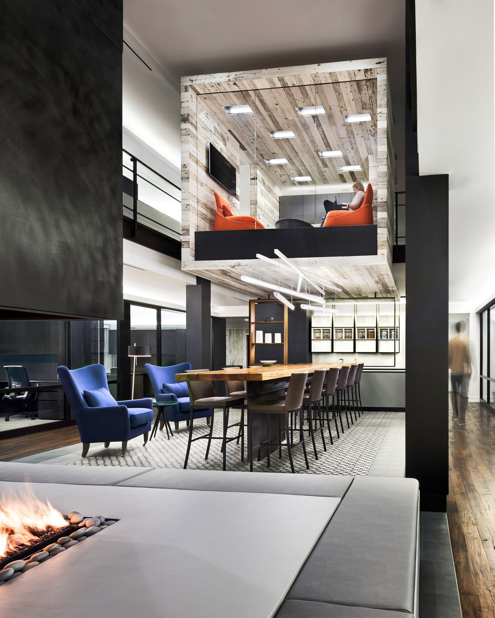Modern open-plan office space utilizing a monochromatic palette with black accents. Central to the design is a suspended wooden box meeting area with glass sides, furnished with burnt orange armchairs. Below, a seamless transition connects a cozy fireside lounge to a communal workspace with deep blue chairs.