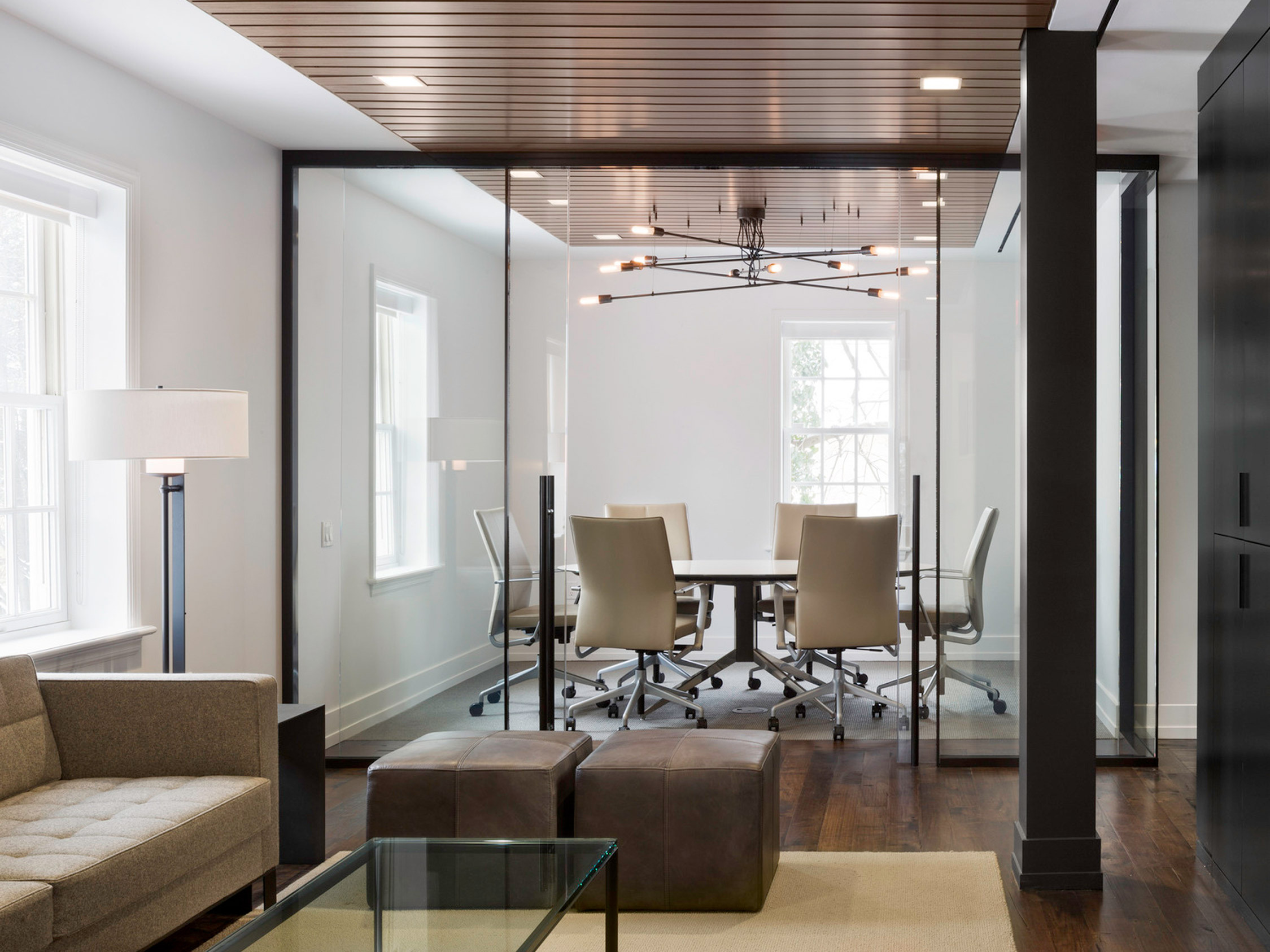 Modern conference room with a sleek, minimalist aesthetic. Features warm wood slat ceiling accents, recessed lighting, and a suspended spider chandelier. Floor-to-ceiling windows provide ample natural light, complementing the neutral color palette and clean lines of the furnishings.