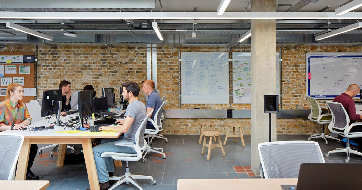 Open-plan workspace with exposed ductwork and brick walls, filled with collaborative clusters of desks and whiteboards, bustling with team interaction and brainstorming.