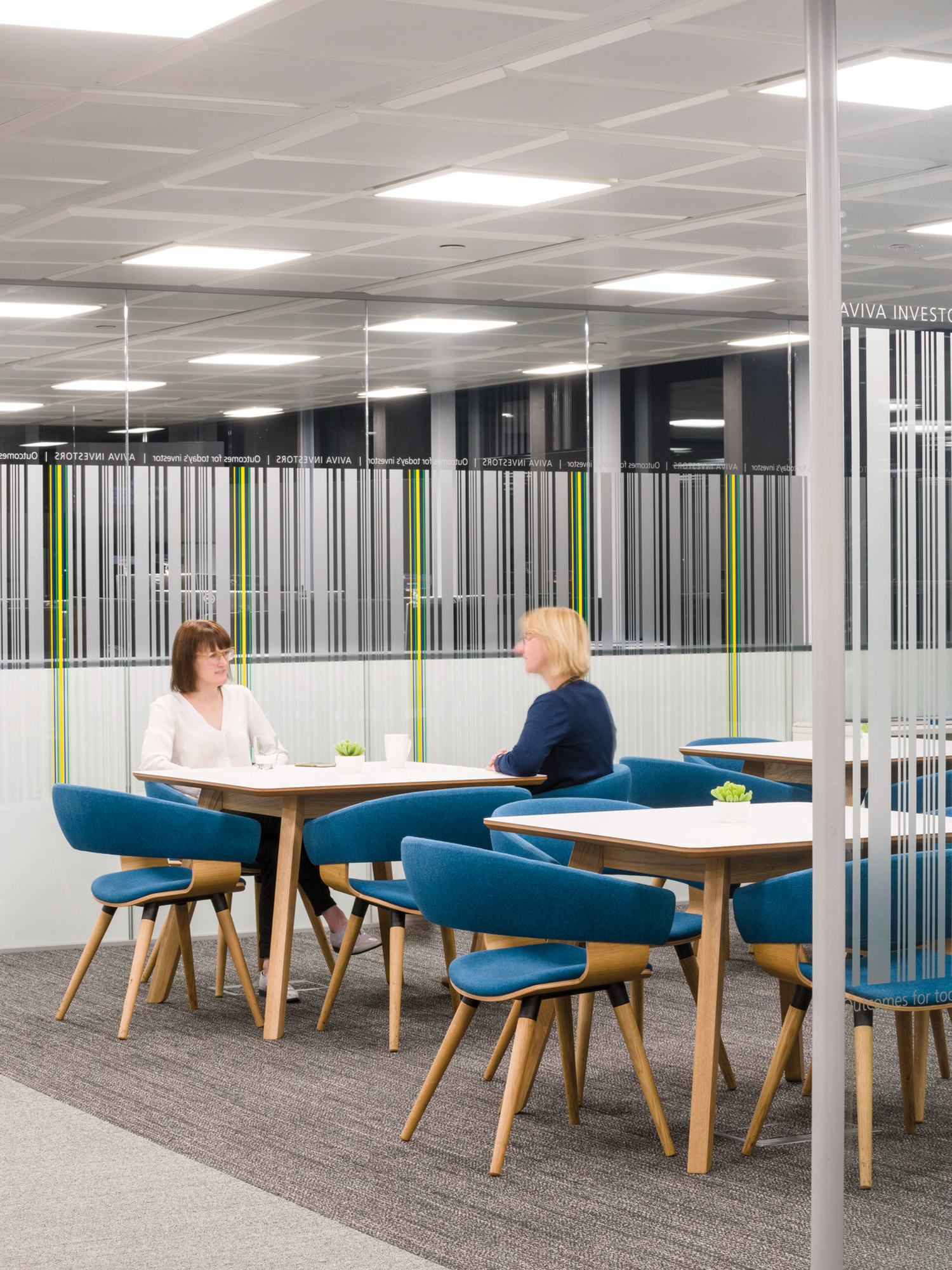 Modern office meeting area with a Scandinavian-inspired design, featuring sleek wooden furniture with blue upholstered chairs, set against a backdrop of translucent dividers with vibrant vertical accents.