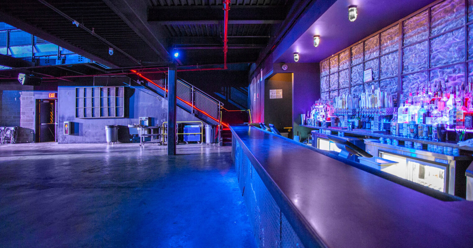 Spacious nightclub interior with vibrant blue lighting accentuating the transparent liquor display. A sleek, black-topped bar runs parallel to an open dance floor, and red pipes overhead add a burst of contrasting color to the industrial-chic ambiance.