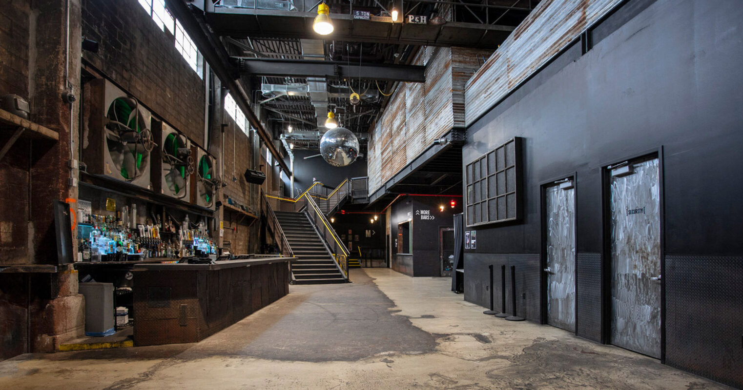 Spacious industrial-style venue featuring exposed brick walls, steel beams, and a bar area with eclectic bottle display. A staircase with metal railings ascends to a mezzanine level, underlining the venue’s multi-functional layout and urban aesthetic.