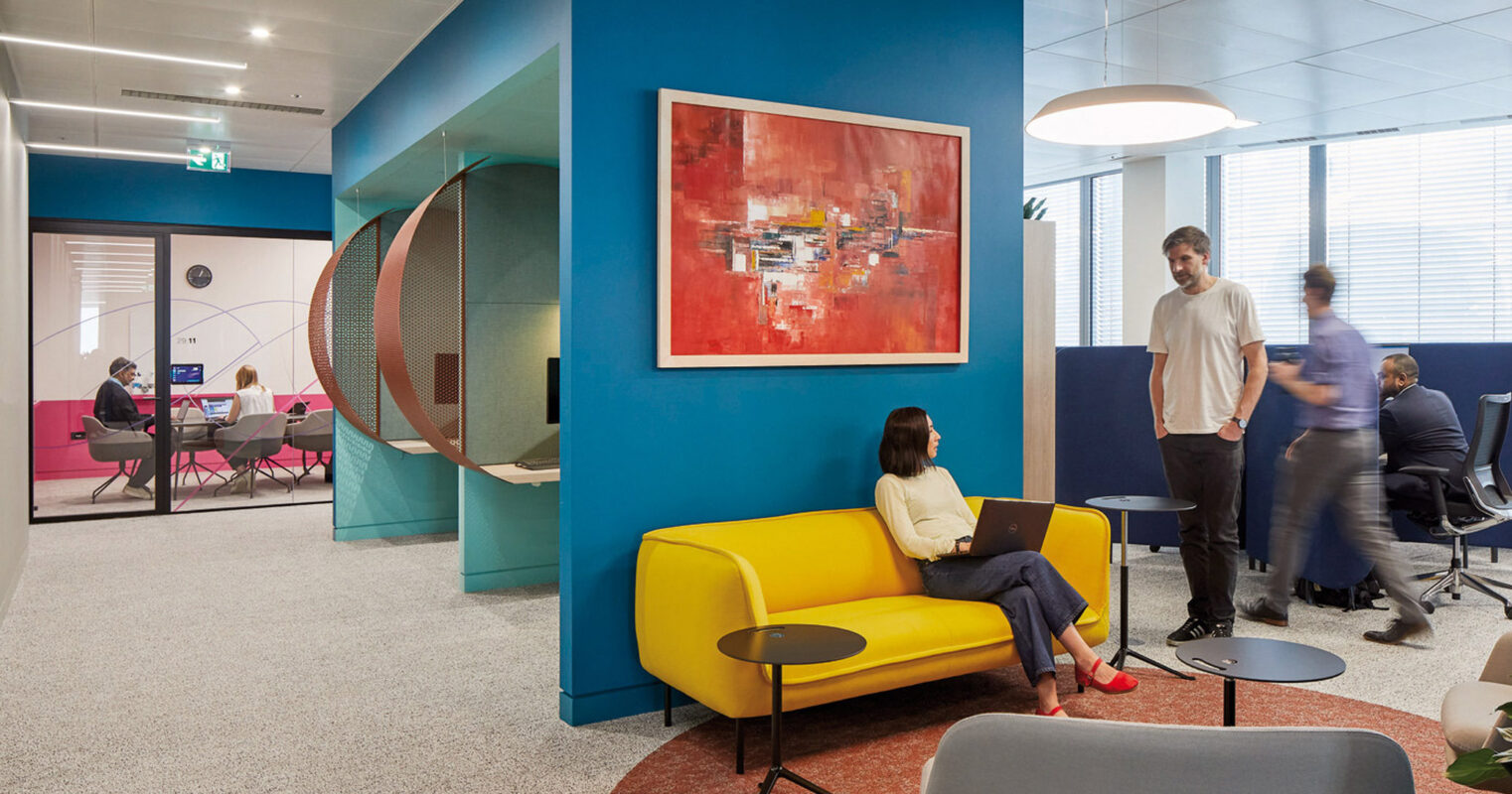 Vibrant modern office space featuring an eclectic mix of geometric shapes and bold colors. A circular cutout connects two areas, with employees engaging nearby. Artwork on blue accent wall complements a yellow sofa, while a gray armchair offers contrast, embodying a dynamic and creative work environment.