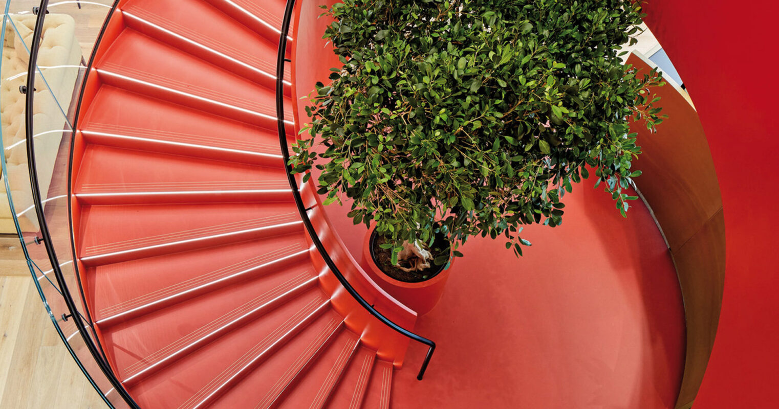 Spiral staircase with striking red walls encircled by metallic balusters. Below, a single figure walks, offering scale, beside a central green potted tree creating a natural contrast to the bold architectural lines.
