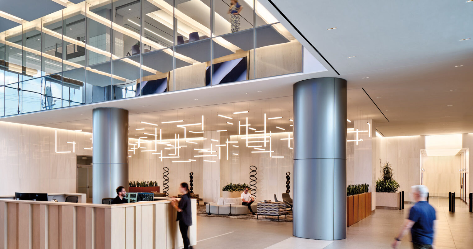 A sleek and modern corporate lobby featuring a reception desk, stylish hanging lights, and multi-level walkways with glass balustrades, with people entering and moving about the space.
