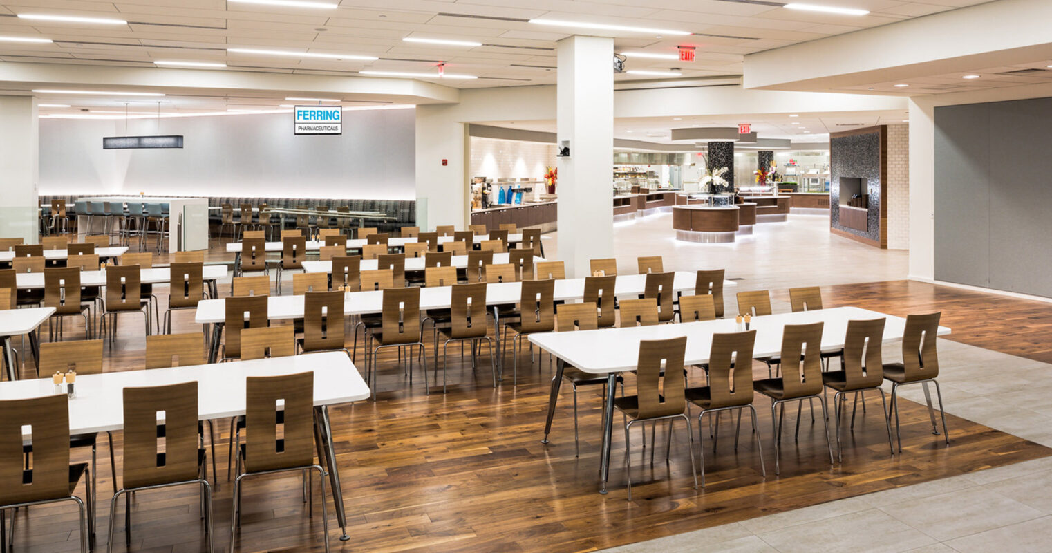 Spacious, modern cafeteria interior with rows of parallel brown tables and chairs on polished wooden flooring. White ceiling with square red accents and recessed lighting complements the open, airy atmosphere. A serving area with counters is visible in the background.