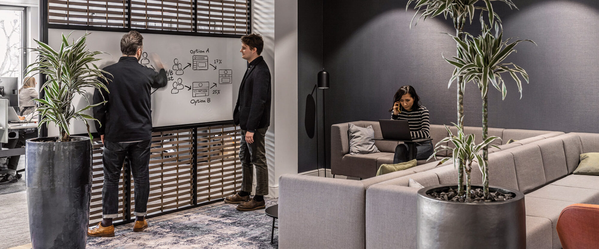 Contemporary office lounge with two individuals discussing a whiteboard, another seated on a gray modular sofa. Slatted wooden screens, lush potted plants, and a patterned area rug create a warm, inviting workspace.