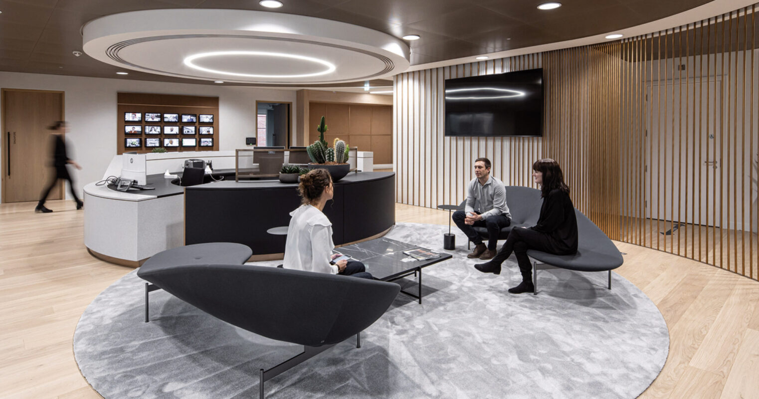 Modern office lounge featuring curved design elements, casual black seating, and a central gray area rug. Wooden slats flank one side, enhancing the room's warm yet sophisticated aesthetic. A circular reception desk sits in the back with ambient lighting overhead.