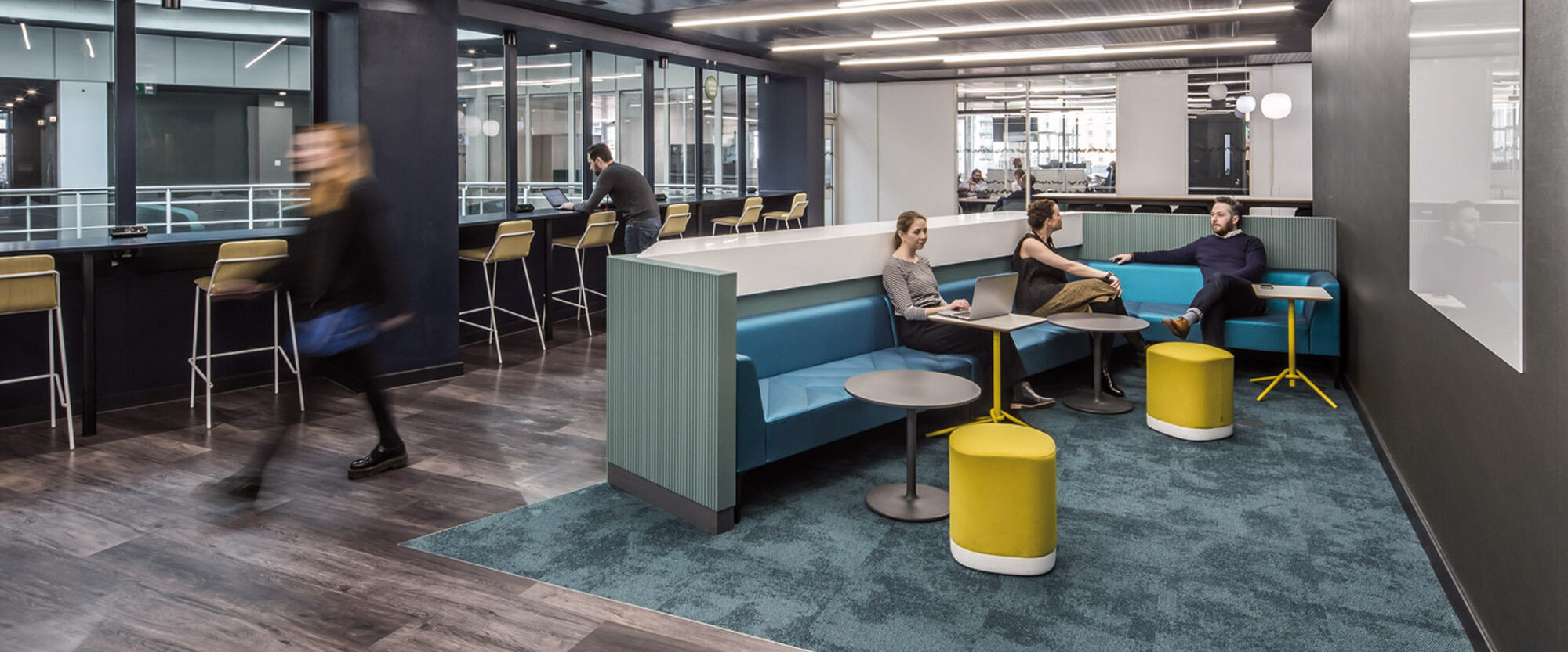 Modern office break area with sleek, linear lighting overhead, teal carpeting, and an eclectic mix of seating options including booths, stools, and pouffes. The space is accented by pops of bold yellow and cool blue tones, fostering a dynamic and collaborative environment.