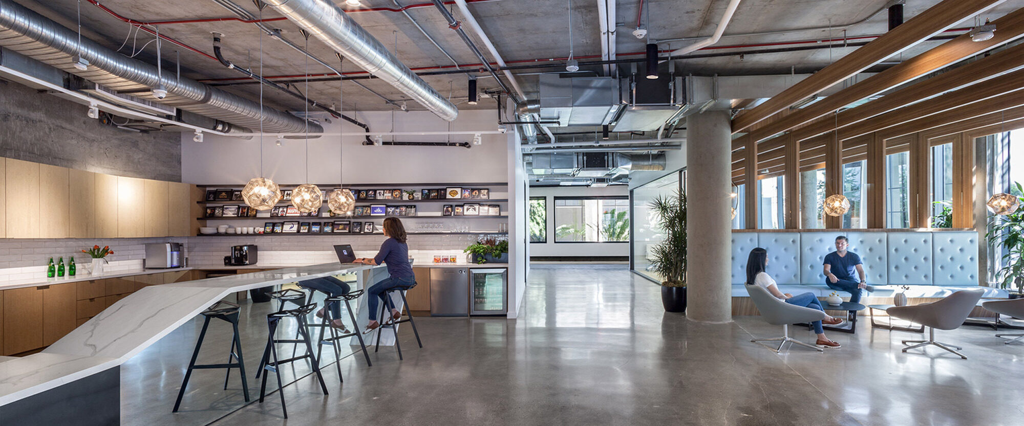 Open-concept office space featuring exposed ceiling beams, polished concrete floors, and a kitchenette with wooden accents. Pendant lighting illuminates the bar seating area, while employees engage in casual conversations in a lounge setting with modern chairs and coffee tables.
